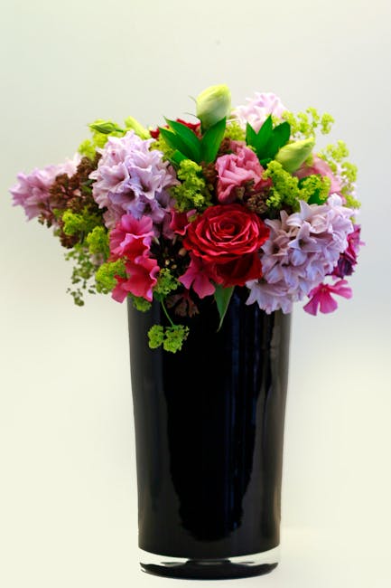 A tall, elegant black glass vase holding a vibrant floral arrangement featuring a large, deep red rose at the center, surrounded by light pink and purple hydrangeas, white and pale green buds, and lus