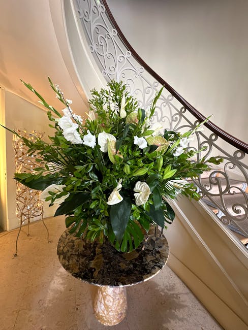 A large floral arrangement featuring predominantly white flowers, including lilies and anthuriums, complemented by lush green foliage with long, pointed leaves and broad, glossy leaves. The bouquet is