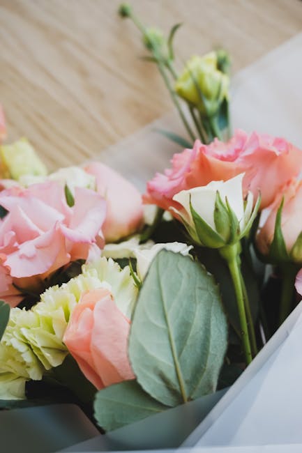A close-up of a floral arrangement featuring light pink and cream eustoma (lisianthus) flowers with ruffled petals, accompanied by green foliage and leaves. The bouquet is wrapped in transparent cello