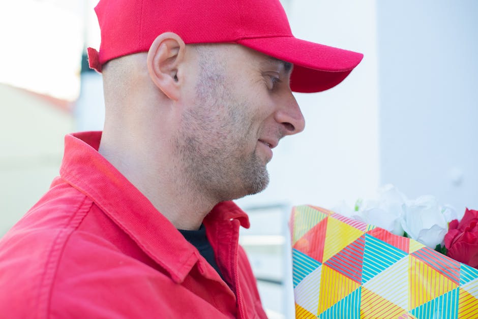 A delivery person dressed in a red cap and red jacket is shown holding a floral gift in a colorful, geometric-patterned wrapping paper with shades of yellow, red, blue, and green. The person is smilin